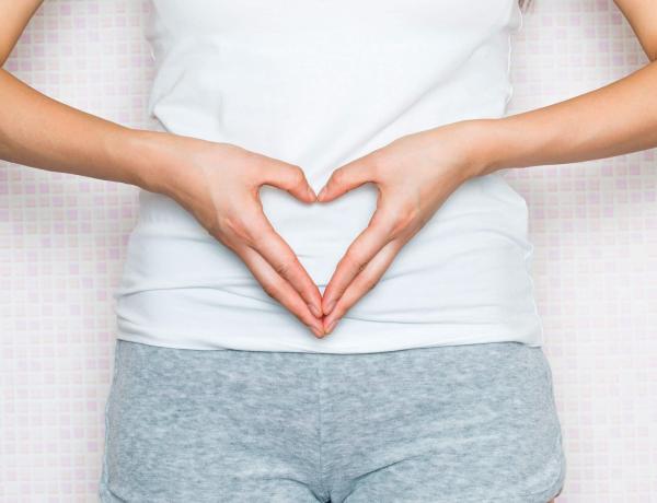 Eine Frau steht vor einer rosa-gekachelten Wand und hat ein weißes Top und graue Shorts an. Sie hält ihre Hände in Herzform vor ihren Bauch.