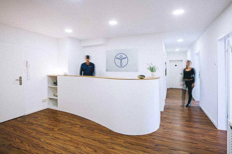 Links im Eck ist die Eingangstür, in der Mitte eine halbrunde Rezeption, darüber hängt das Logo der Praxis. Die Menschen sehen verschwommen aus.